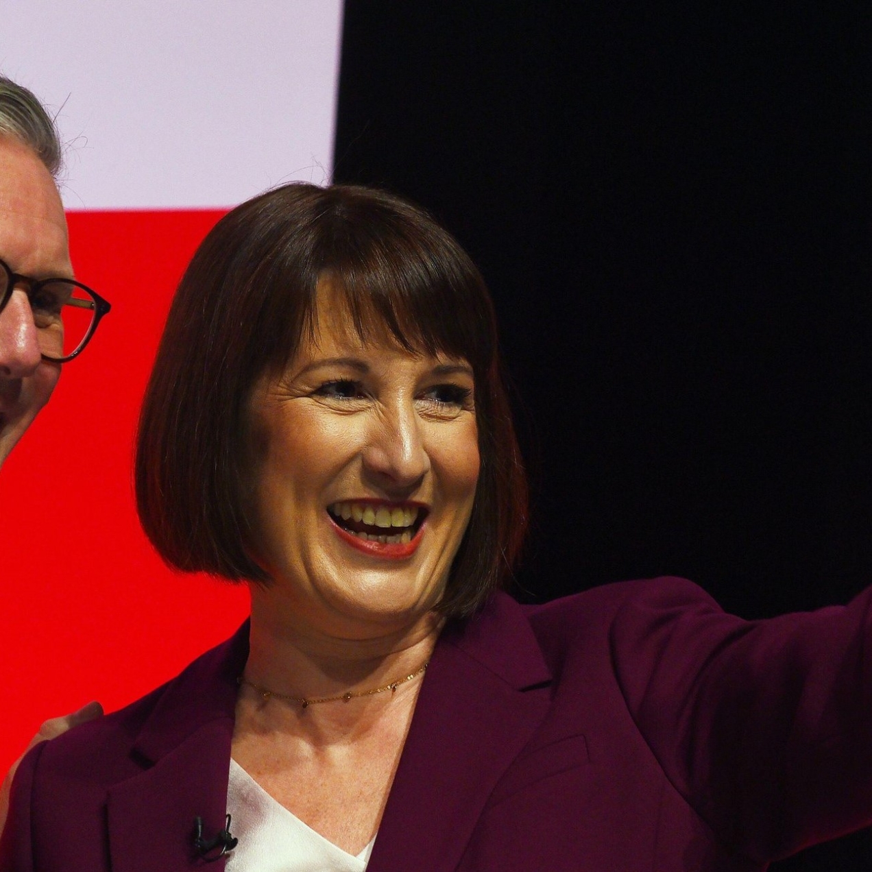 Kerr Starmer and Rachel Reeves smiling at the Labour Party conference. Starmer is a white man with grey hair, wearing a dark blue suit and glasses. Reeves is a white woman with short dark hair wearing a burgundy jacket over a white top, she is waving